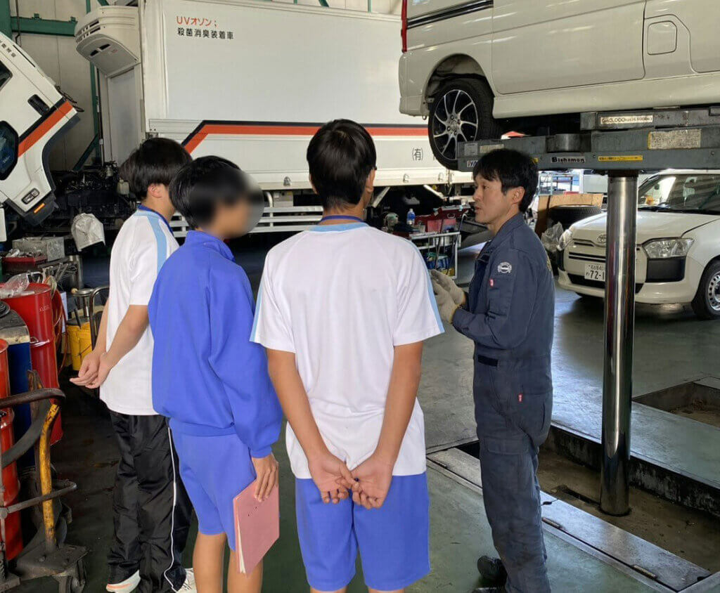 中部第一自工の整備工場で車両整備について学ぶ蟹江中学校の生徒。リフトで上がった車の下で整備士が説明している職場体験の様子
