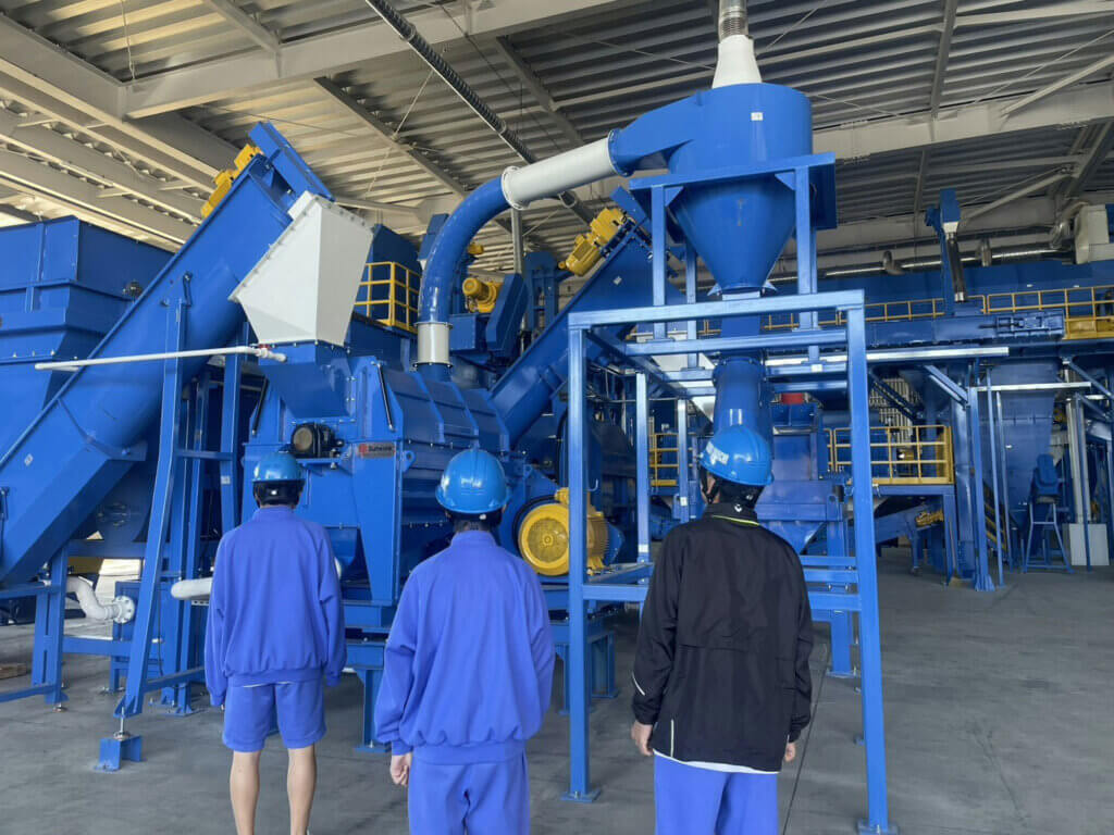 蟹江中学校の生徒がエコロジスティクスセンターの大型リサイクル設備を見学。青いヘルメットを着用した生徒が最新の破砕・選別機を見上げている