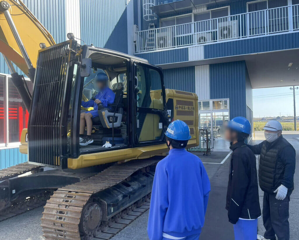 蟹江中学校の生徒がCAT油圧ショベルの運転席に乗り込む体験。青いヘルメットを着用した生徒が重機の操縦席で貴重な体験をしている