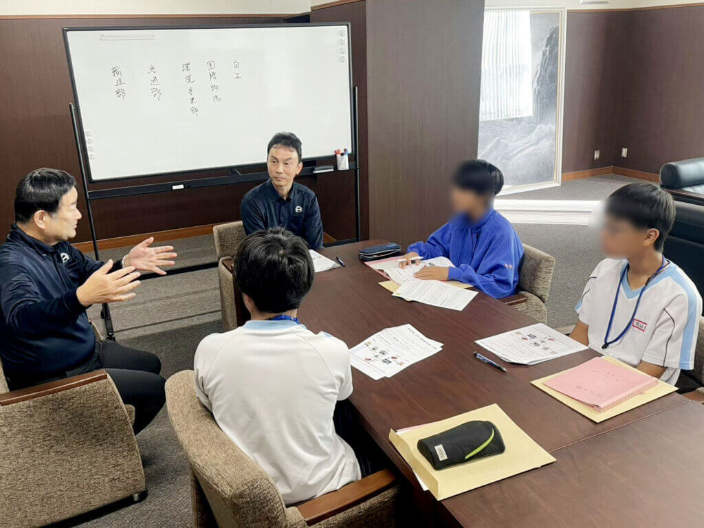 中部第一輸送のリサイクル現場で社員がリサイクル素材について説明。蟹江中学校の生徒3名が真剣に話を聞いている職場体験の様子