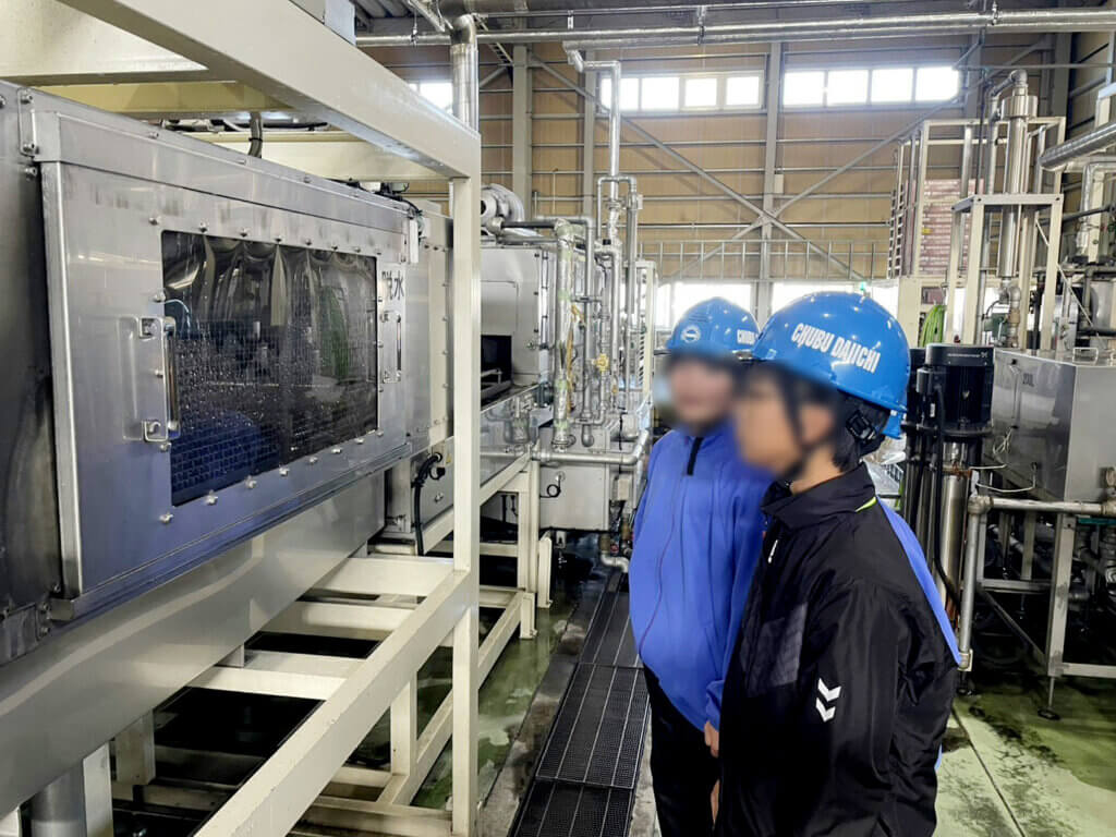 蟹江中学校の生徒がパレット洗浄施設を見学。中部第一輸送のパレット洗浄機の前でヘルメットを着用した生徒が説明を聞いている