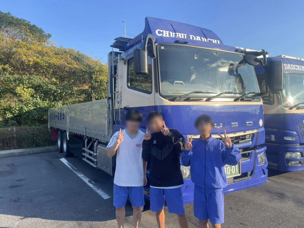 中部第一輸送の大型トラックの前でピースサインをする蟹江中学校の生徒3名。青空の下、FUSOトラックと一緒に記念撮影