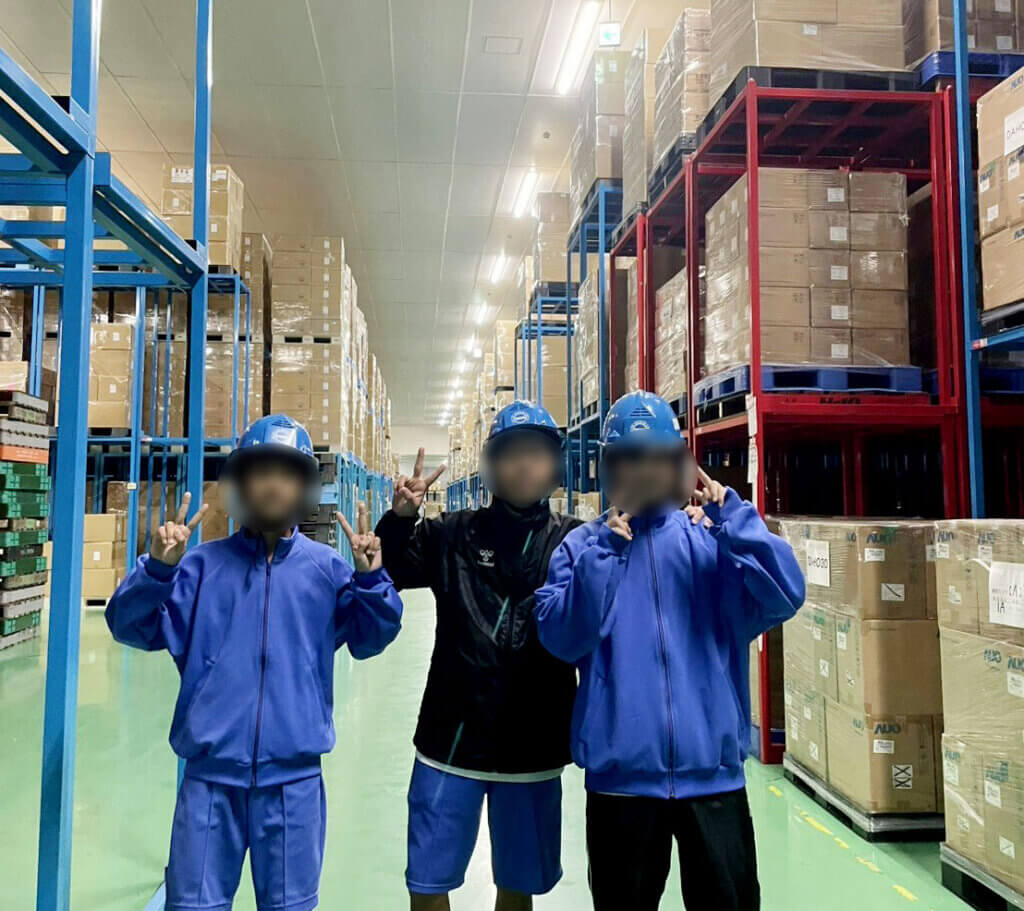 中部第一輸送の倉庫内でピースサインをする蟹江中学校の生徒3名。青いヘルメットを着用し、高い棚に囲まれた倉庫で笑顔の集合写真