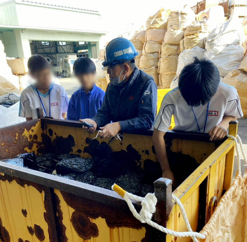 中部第一輸送のリサイクル現場で社員がリサイクル素材について説明。蟹江中学校の生徒3名が真剣に話を聞いている職場体験の様子