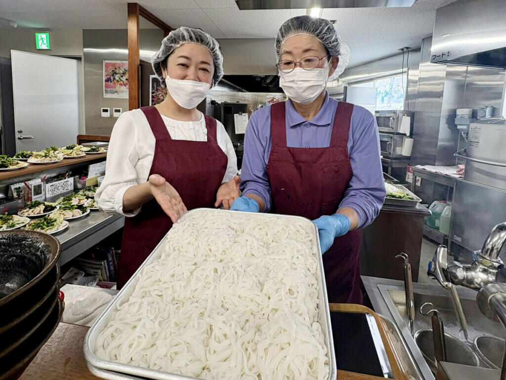 京都から来た小平先生と中部第一輸送の食堂スタッフがベトナム料理フォーの麺を持って笑顔。料理研修の様子